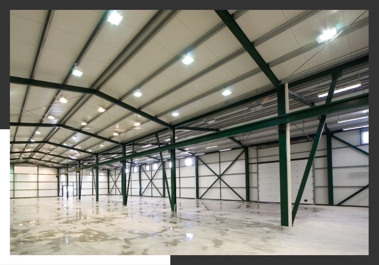 The wide, empty interior of a large, modern warehouse, showing the green steel support columns and roof trusses.
