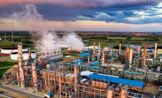 Aerial view of a sprawling petrochemical or oil refinery complex with interconnected pipes, storage tanks, and towers, emitting steam against a vivid sunset or stormy sky over a green landscape.
