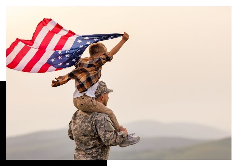 military member holding child on shoulders while he flies the American flag