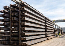 A variety of heavy structural steel beams and channels are organized on tall racks in a brightly lit industrial fabrication yard under natural daylight.